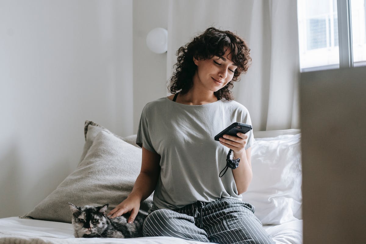 Smiling young ethnic female messaging on smartphone and petting cat on bed

