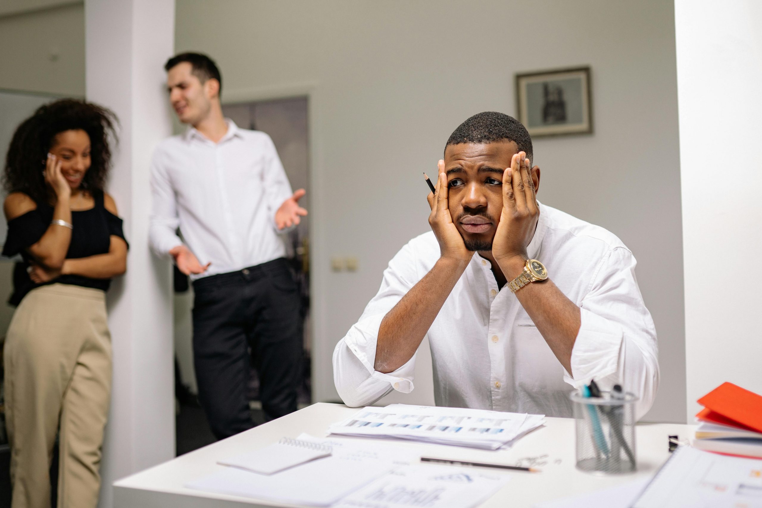stressed employee sitting at his desk while two other employees happily chat behind him